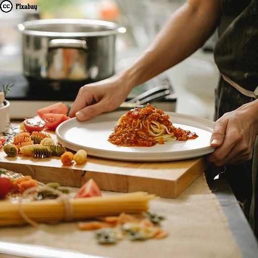 Eine Person richtet in einer K&uuml;che frisch gekochte Spaghetti mit Tomatensoße auf einem Teller an. Auf der Arbeitsfl&auml;che liegen verschiedene Zutaten und Nudeln bereit.