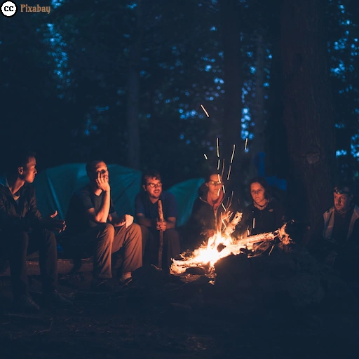 Mehrere Menschen sitzen abends gemeinsam an einem knisternden Lagerfeuer auf einem Campingplatz im Wald.
