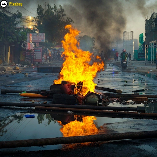Eine brennende Barrikade auf einer verw&uuml;steten Straße, w&auml;hrend dichter Rauch &uuml;ber den Ort zieht.