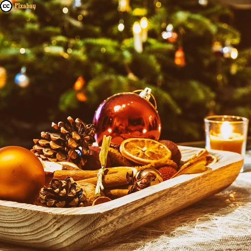 Eine hölzerne Dekoschale mit Weihnachtskugeln, Tannenzapfen, Zimtstangen und getrockneten Orangenscheiben steht vor einem festlich geschmückten Weihnachtsbaum. Mehrere Kerzen tauchen die Szene in warmes Licht.